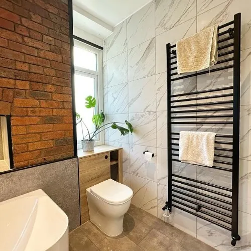 An urban-chic bathroom blending modern aesthetics with a touch of rustic charm. Exposed brickwork complements the cool gray marbled tiles, creating a delightful contrast. A tall black towel rack stands out, holding plush white towels. A wooden vanity holds a potted plant by the window, bringing a breath of fresh greenery into the space. The gleam of the toilet and bathtub, along with the natural light filtering through, adds a serene touch to the room