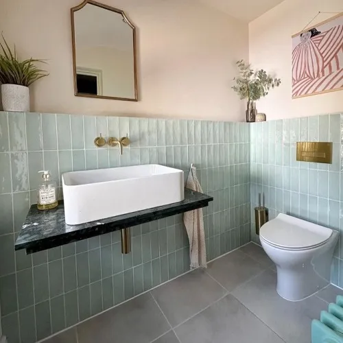 "A chic and modern bathroom showcasing mint-green tiled walls paired with a sleek white rectangular basin atop a dark green marble countertop. The space is accented with gold fixtures, a contemporary mirror, and pops of pastel décor including a playful pink-striped wall hanging. Lush potted plants and a soft hand towel add cozy finishing touches