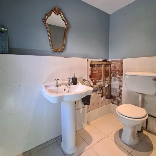 Bathroom with pale blue walls, featuring a white pedestal sink and toilet, complemented by a vintage gold-framed mirror. One section of the wall reveals exposed brickwork, indicating ongoing renovations