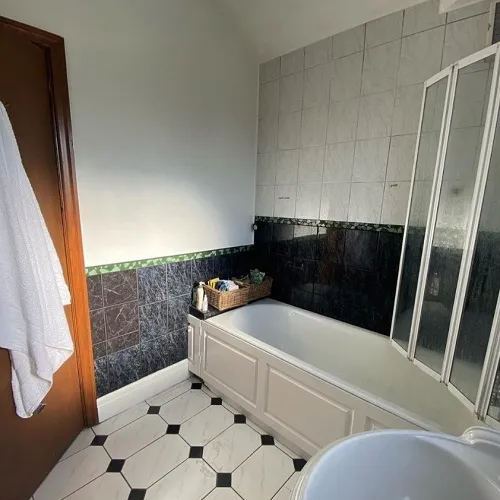 A neat bathroom featuring a mix of monochromatic tiles; from the glossy dark green and gray on the walls to the geometric white and black pattern on the floor. A white bathtub with a sliding glass door is nestled against the tiled wall, equipped with a shelf holding toiletries. Warm sunlight filters in, casting a glow on the room. A fluffy white towel hangs conveniently close on the wooden door to the left