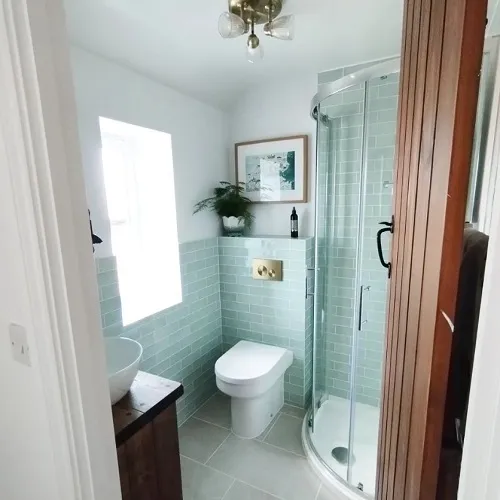 Bathroom featuring teal subway tiles, a glass-enclosed shower, a modern bowl-style sink atop a wooden vanity, and a white toilet. A window lets in natural light, and there's a framed artwork on the wall.