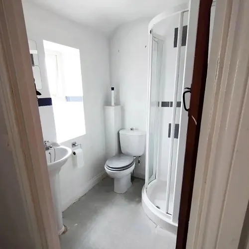 Compact bathroom with a curved corner shower unit, pedestal sink, and a toilet. The space is painted in white and has a window for natural light. 