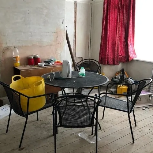A room in the midst of transformation, telling a story of renovation and creativity. A circular table stands at the center, surrounded by wire-frame chairs and littered with various tools and items, from paint cans to a vacuum. The walls are bare, waiting for a fresh coat, while a lone crimson curtain flutters by the window. Evidence of hard work is everywhere, hinting at the potential beauty this space will soon embrace.