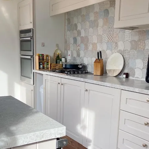 Modern kitchen space with white cabinets and a patterned splashback. Features a built-in oven and a countertop stove. The counter hosts cooking essentials and some groceries.