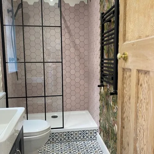 A trendy bathroom space showcasing a blend of vintage and modern design elements. Honeycomb pink tiles envelop the shower area, contrasted by a clear glass partition with black framing. A white sink sits beside the shower, while the floor boasts an intricate monochrome pattern. The room's aesthetic is completed by a tropical-themed wallpaper section and a wooden door with classic detailing. A black heated towel rack is a chic and practical touch