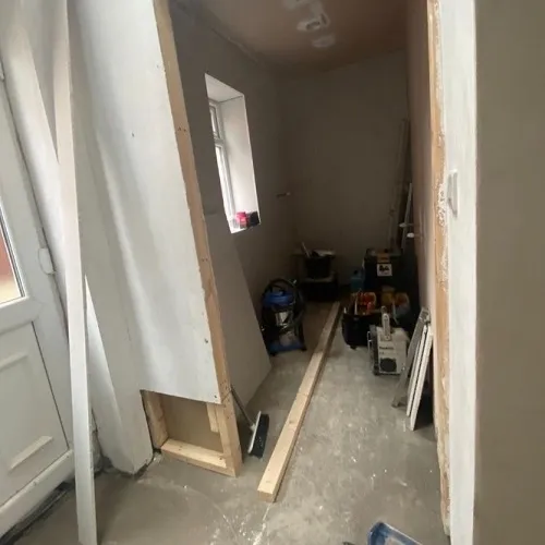 undergoing renovations with visible wooden framing on one side and freshly plastered walls. The floor is scattered with construction materials, tools, and a vacuum cleaner, highlighting the active work in progress. A small window lets in a glimpse of natural light to the space.
