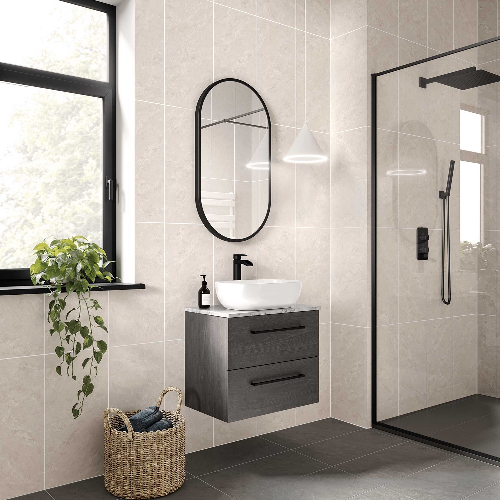 A modern bathroom with beige textured wall tiles and dark grey floor tiles. The room features a floating vanity with a wood finish, topped with a white vessel sink, next to a large, oval-shaped mirror with a black frame. A clear glass shower enclosure with a black shower system is visible to the right. Natural light streams in from a window, and there's a potted  green plant on a black windowsill, adding a touch of nature to the space. A woven basket filled with towels sits on the floor, suggesting a spa-like atmosphere. 