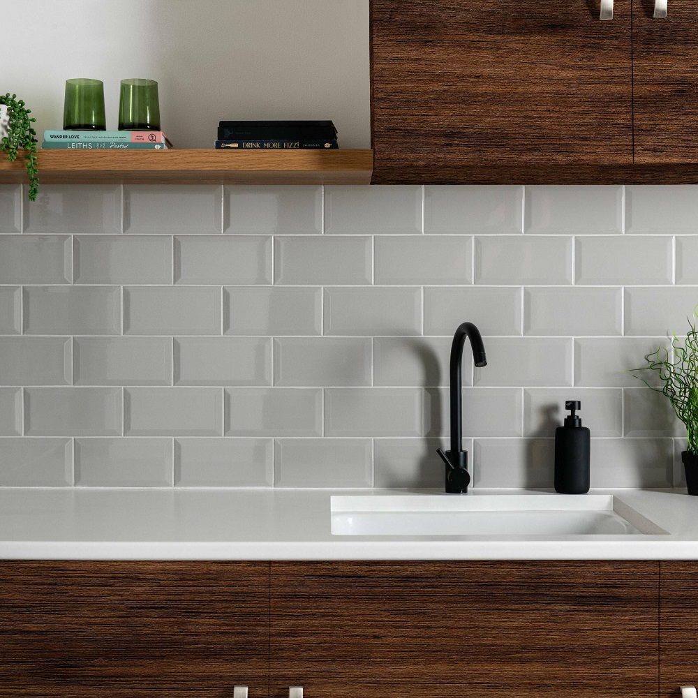 A stylish kitchen featuring light grey metro wall tiles above a white countertop. A dark wood cabinet with modern handles and a black, tall neck faucet enhance the contemporary aesthetic. A wooden shelf above the sink displays two green glass jars, a black book, and a small green plant, adding a touch of colour and life to the space. The design is clean and minimalist, with a focus on functionality and modern lines. 