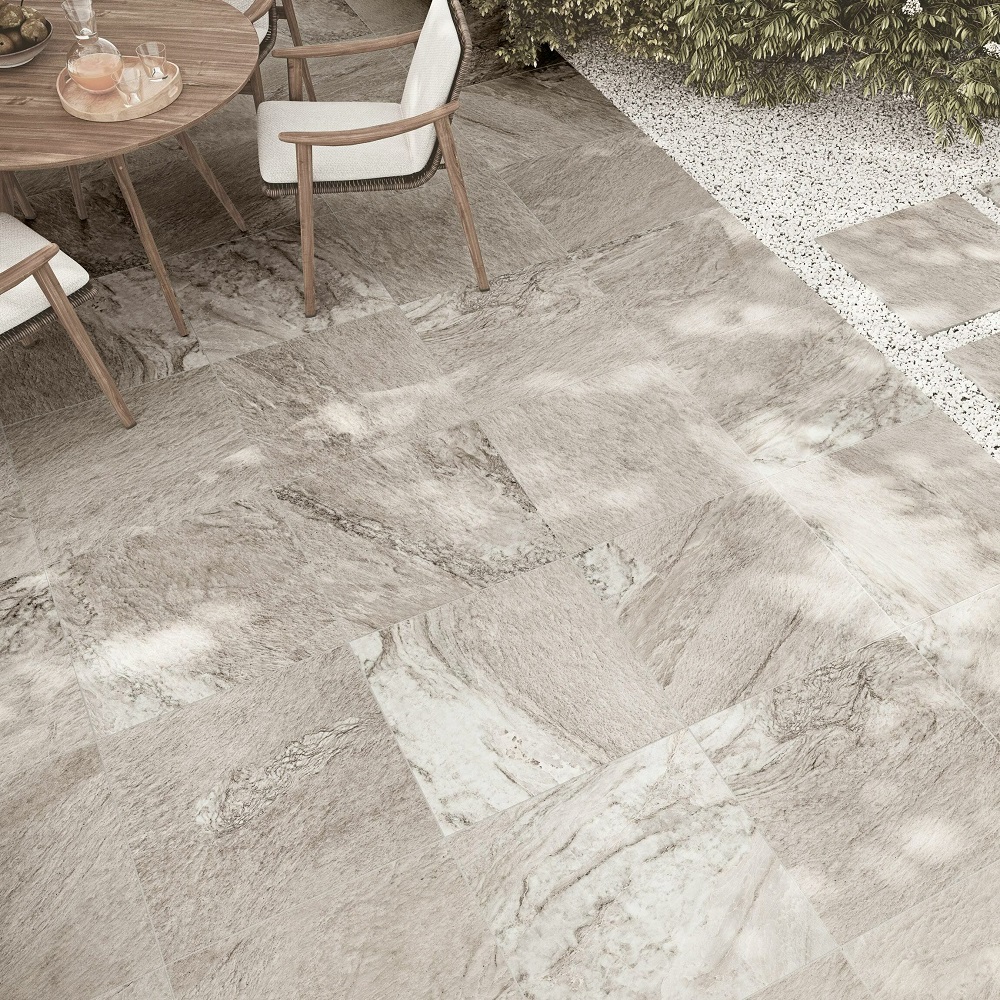 A contemporary patio featuring large, porcelain tiles with varied greige tones and a natural stone finish, harmoniously blending with the outdoor wooden furniture and gravel border. 