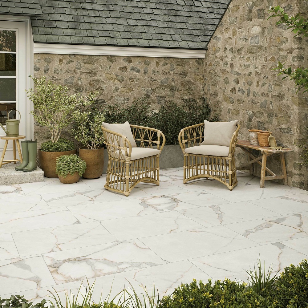 An inviting garden patio area featuring elegant Carrara gold marble effect porcelain tiles, paired with natural rattan chairs and soft beige cushions. Potted olive trees and lush shrubs add to the serene ambiance, set against a rustic stone wall. 