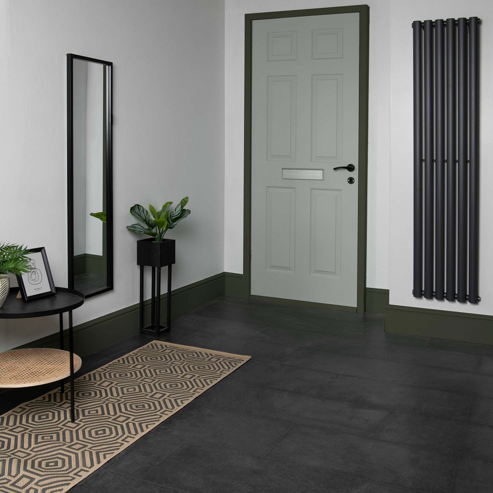 A contemporary hallway featuring concrete effect anthracite tiles laid in a clean, straight pattern. The monochrome palette is complemented by a geometric patterned beige and black runner rug, a slender black framed full length mirror, and a sleek round side table with a wicker basket underneath. A tall vertical radiator adds a modern touch alongside the olive green panelled door, creating a welcoming yet stylish entryway.
