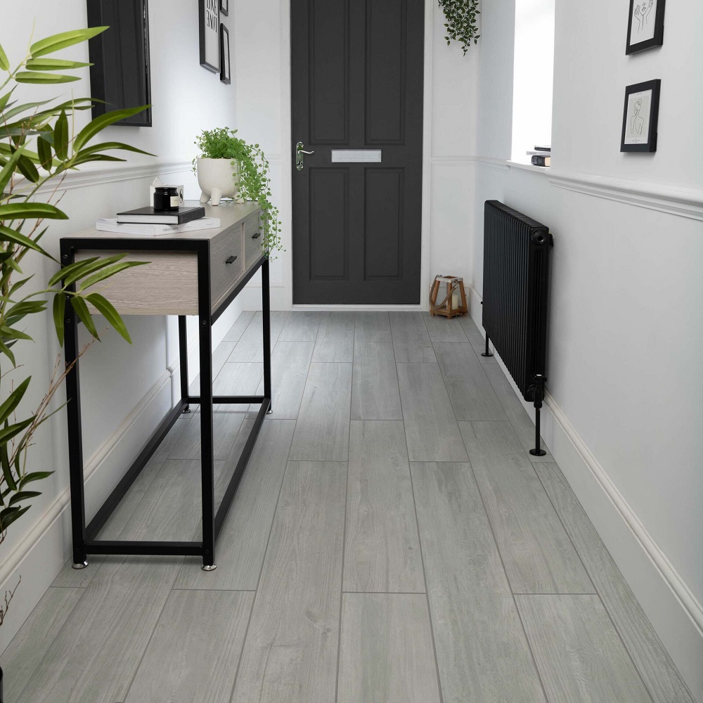 A sleek and modern hallway showcasing rectified matt wood effect tiles in a soft grey, lending a serene and spacious feel to the entryway. The cool tones of the flooring contrast beautifully with the deep charcoal panelled door. The space is styled with a minimalist console table with a black metal frame and light wood top, complemented by small potted plants that add a fresh touch of greenery. White walls with a classic chair rail detail are adorned with an array of chic monochrome framed prints, while a vintage style black radiator adds character to the contemporary setting. 