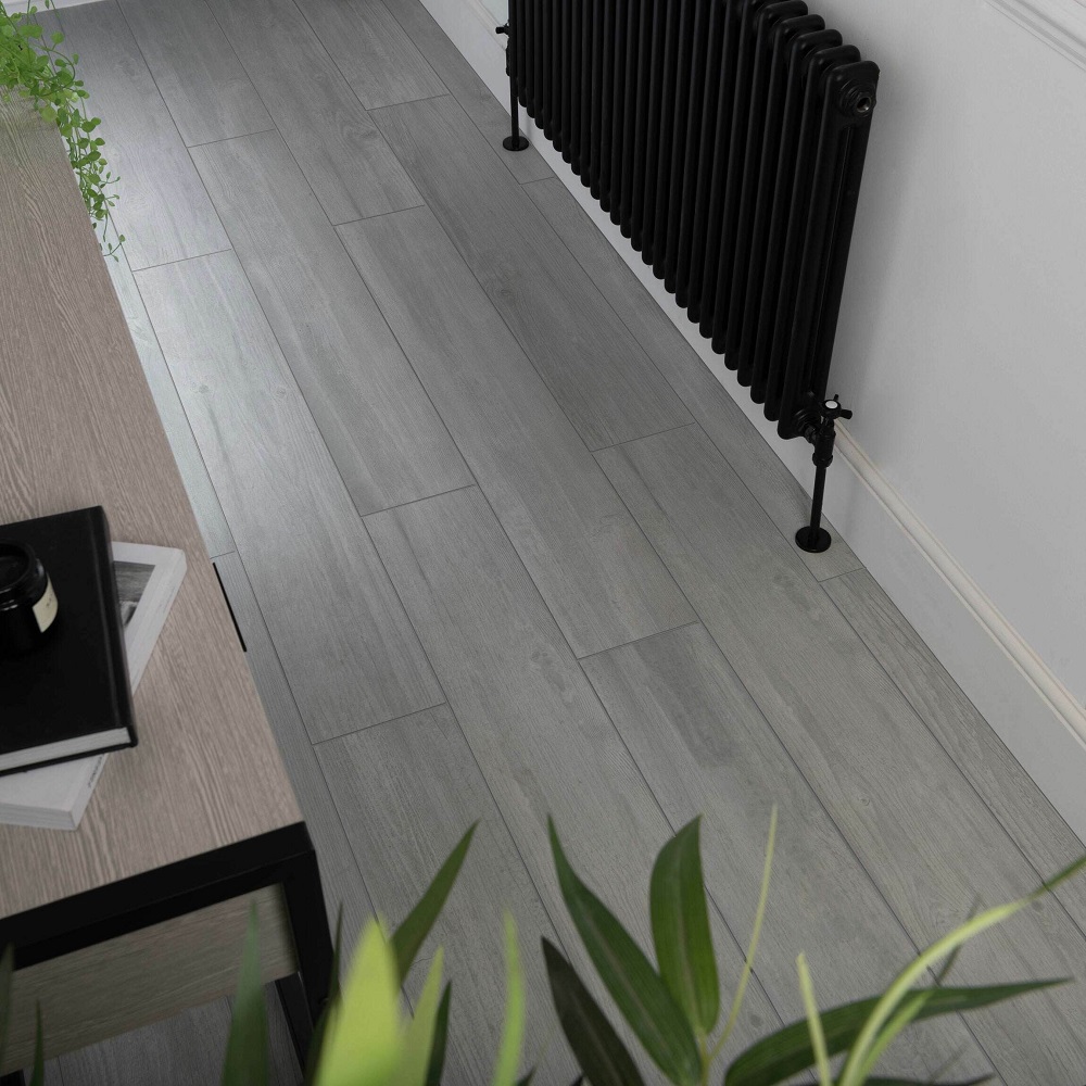 This image captures a contemporary hallway with Copenhagen rectified matt wood effect tiles in a soothing grey tone, laid in a staggered pattern. To the side, a classic black vintage style radiator adds a contrast to the modern flooring. A wooden side table with a black notebook on top and a potted green plant in the foreground introduce natural elements, enhancing the clean and minimalist aesthetic of the space. The walls are a crisp white, complementing the cool tones of the flooring and contributing to the rooms bright and airy feel. 