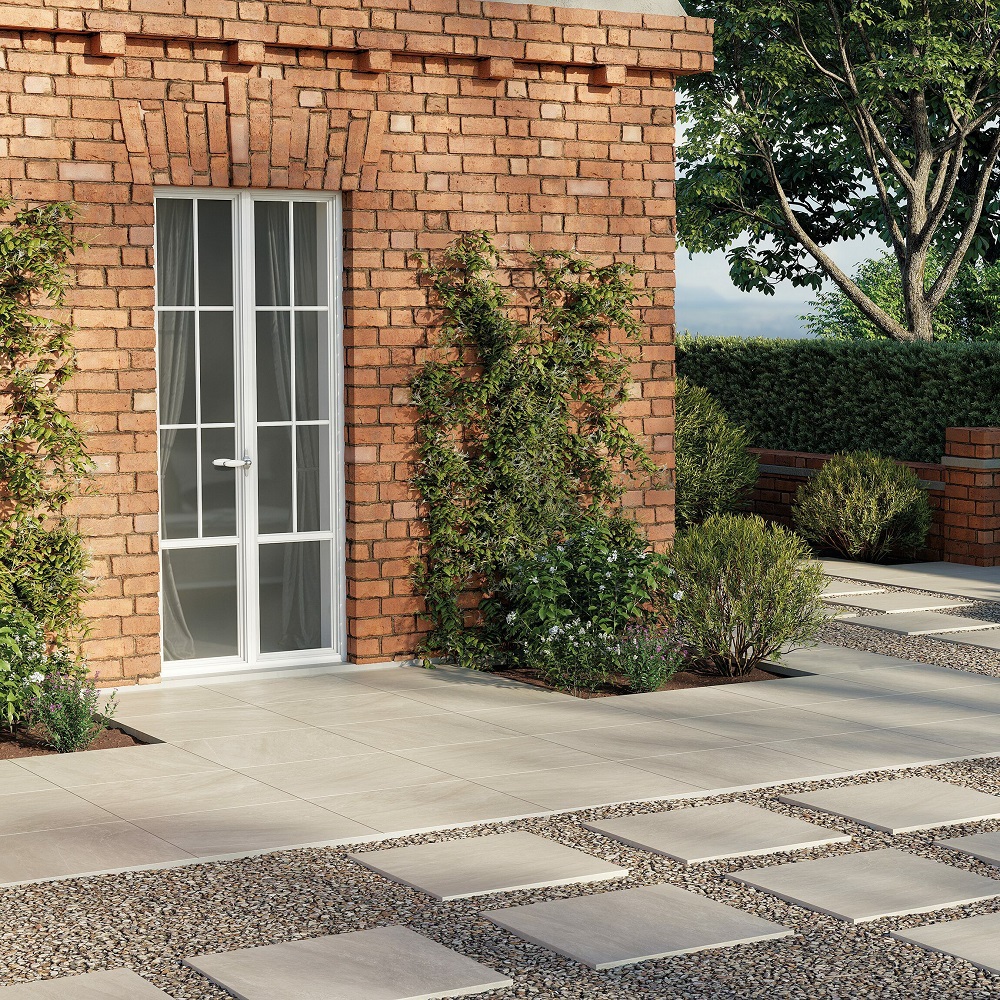 A neatly landscaped path leading to a white door set in a classic red brick wall, complemented by beige porcelain tiles and interspersed with pebbles and square stepping stones. The garden is lush with shrubbery and climbing plants, offering a manicured yet welcoming approach to the home. 