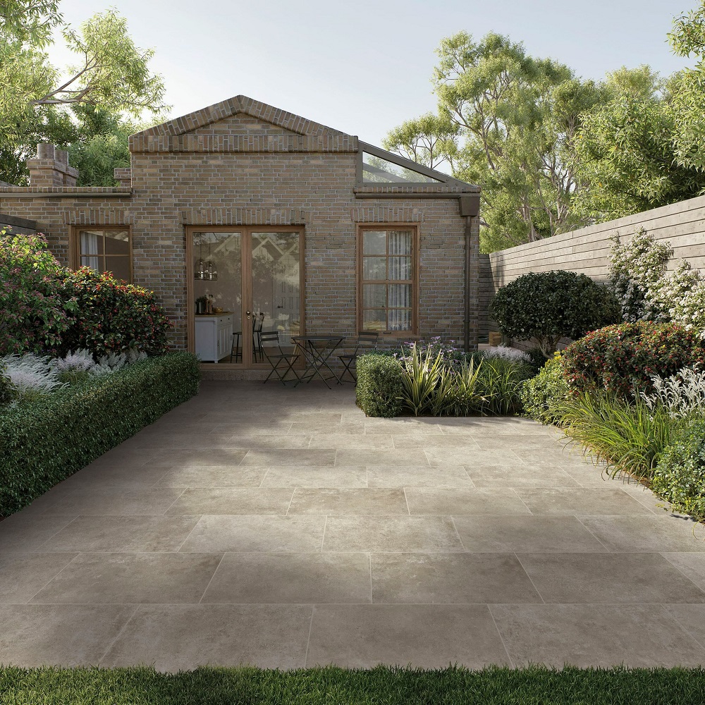 A spacious garden patio with natural concrete-effect matt porcelain tiles, set against an elegant brick conservatory with French doors. The patio is surrounded by lush, manicured hedges and vibrant flowering shrubs, creating a blend of refined structure and natural beauty for outdoor relaxation or entertaining. 
