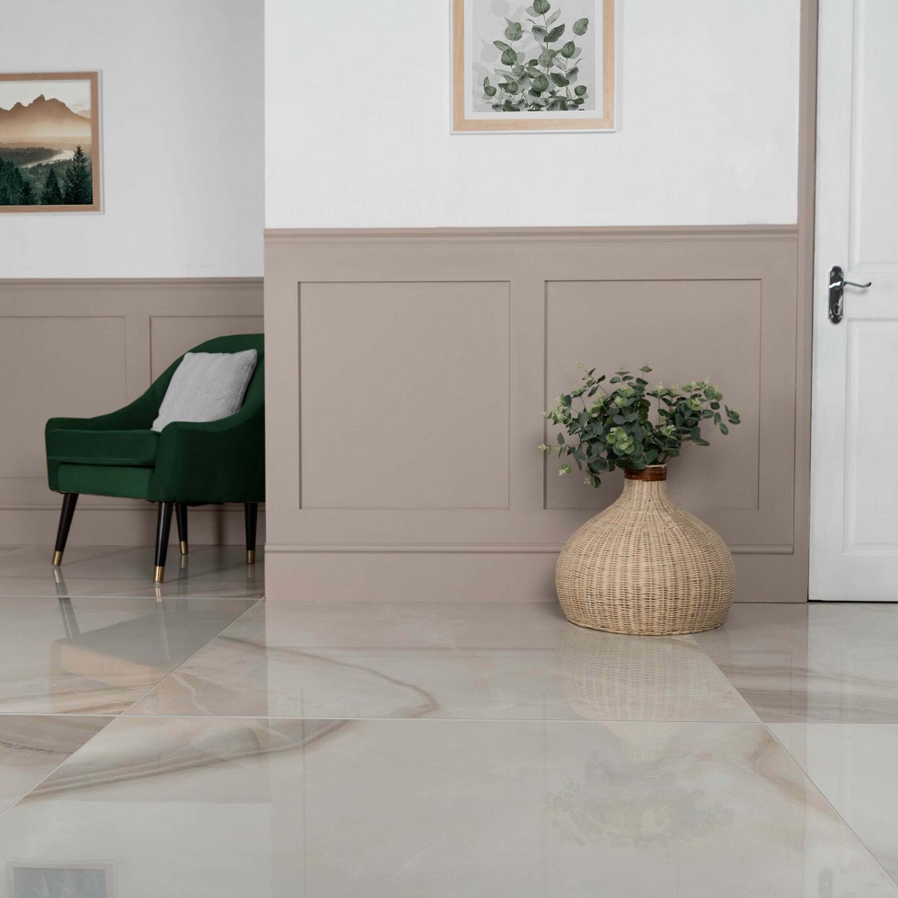 An elegant and airy hallway featuring a large beige polished tile with a delicate onyx marble effect, creating a luxurious and expansive feel. The walls are adorned with classic wainscoting painted in a muted taupe, harmonising with the light floor. A plush emerald green armchair with golden legs adds a pop of colour, while a natural woven vase holding lush greenery brings an organic touch. The scene is completed with framed botanical artwork, enhancing the sophisticated and serene ambiance of the space. 