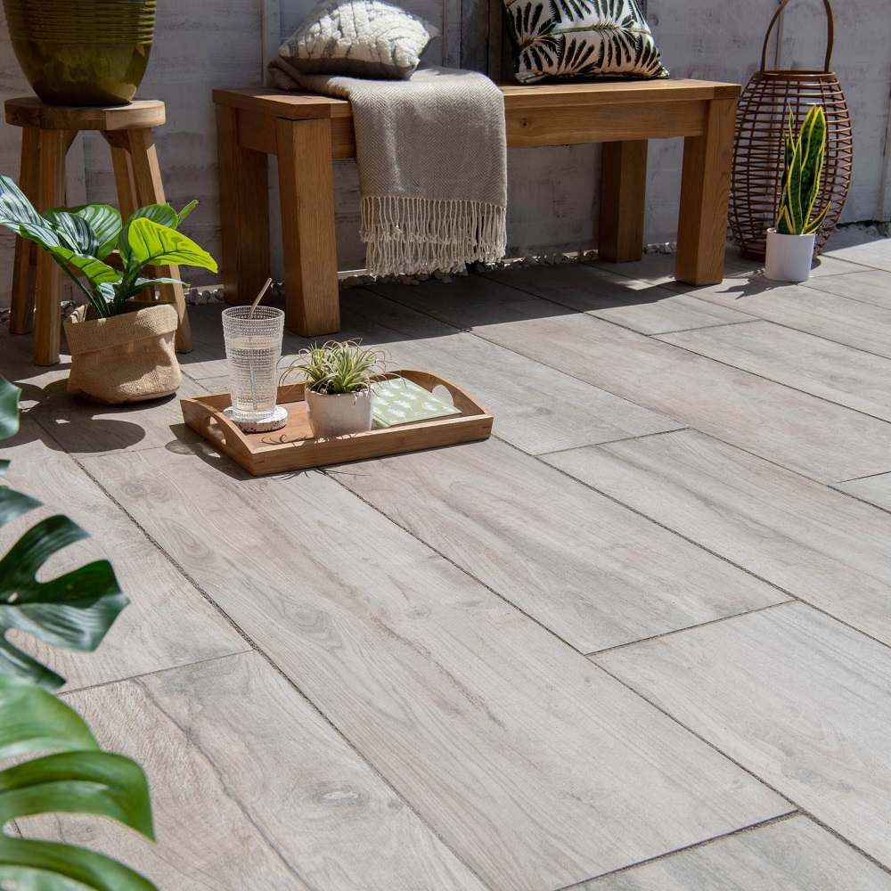 A sun-drenched patio space with light maple-wood effect porcelain tiles, furnished with a wooden bench and stool. The area is accented with greenery, including a potted plant, and a tray with succulents and a refreshing glass of water, invoking a relaxed, outdoor living space.