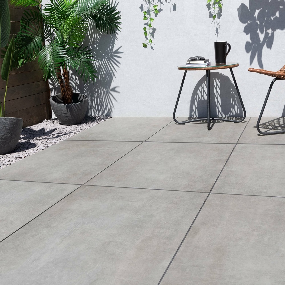 A sleek, modern patio with uniform silver grey porcelain slabs creating a clean and minimalist look, complemented by a side table, a selection of potted tropical plants, and soft shadows from the afternoon sun.