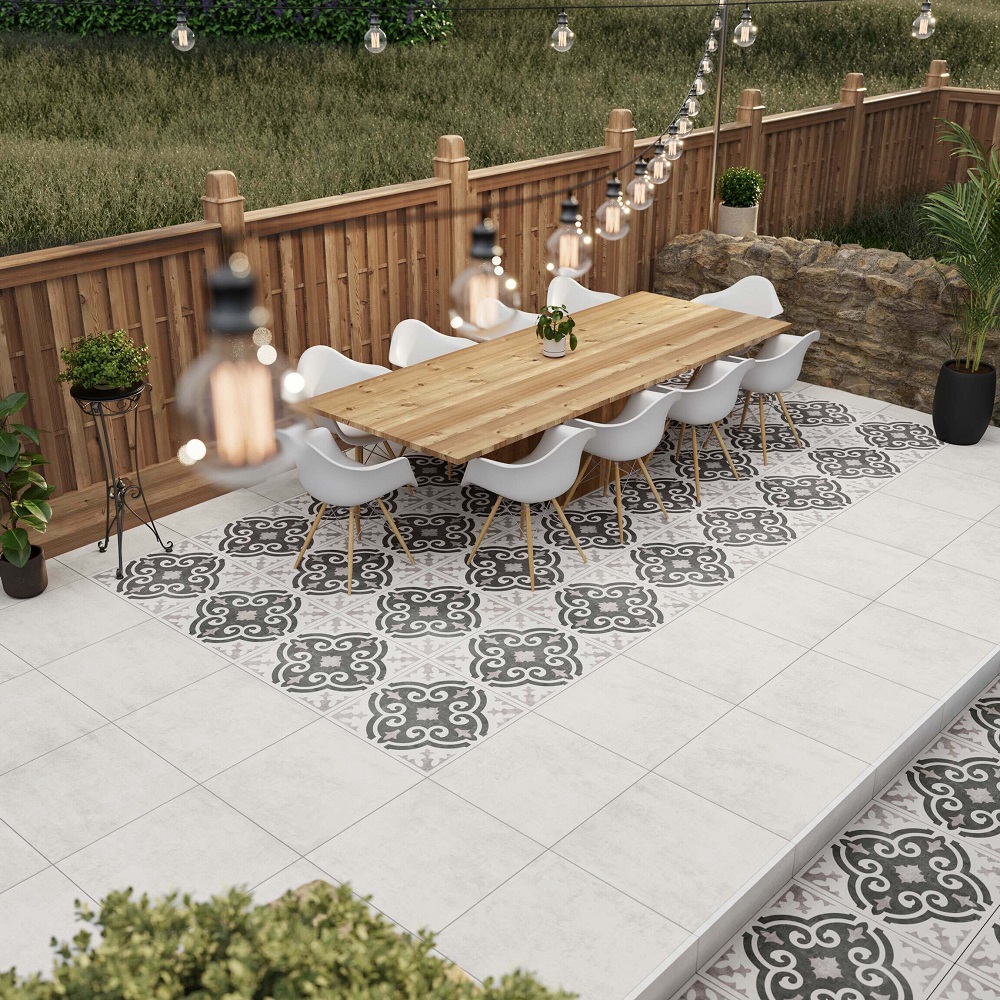 An inviting outdoor dining area with a decorative white and black patterned porcelain tile design under a long wooden table surrounded by white modern chairs. This cosy space is bordered by a wooden fence and adorned with hanging festoon lights, creating a warm and festive atmosphere. 