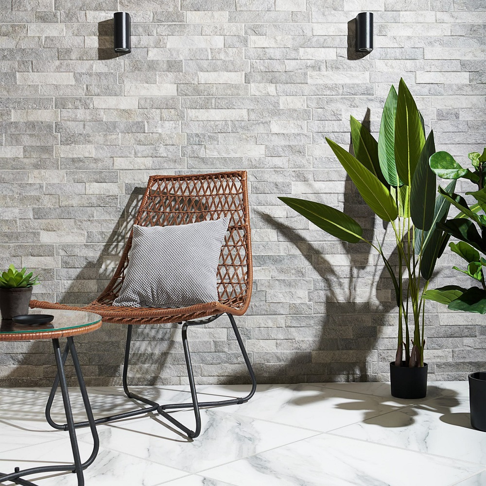 A cosy nook with a textured grey slate effect wall tile backdrop, accented by a modern rattan chair with a comfortable cushion, a side table with a potted succulent, and a tall, vibrant green indoor potted plant adding a touch of nature. 