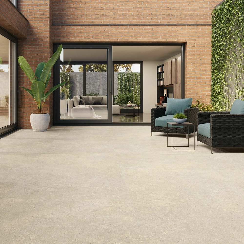 A chic patio area featuring matt stone effect porcelain tiles in a warm cream, complemented by brick walls and contemporary black wicker furniture with teal cushions, The space is accented with a large potted banana plant and has sliding glass doors that provide a view into a modern interior. 