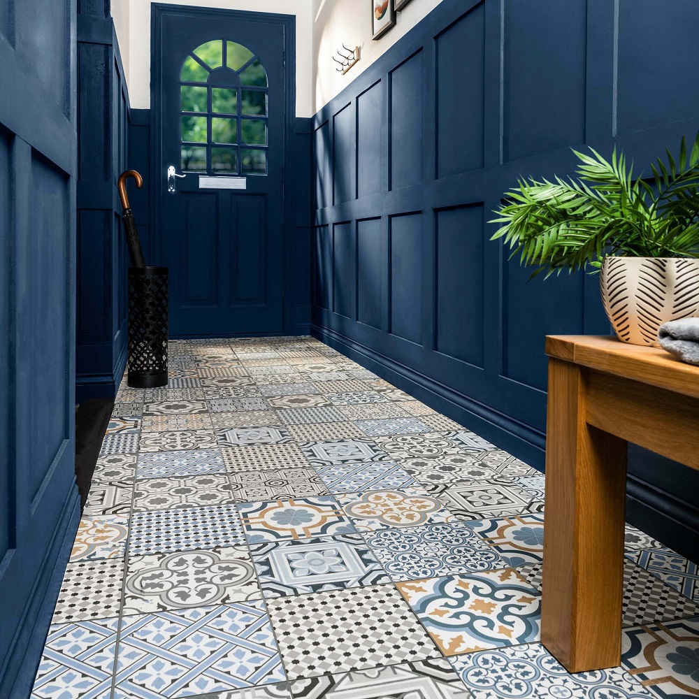 This hallway exudes character with its vibrant patterned porcelain tiles, featuring a medley of blues, greys and creams that combine to form a rich tapestry of designs, reminiscent of traditional encaustic flooring. The bold navy blue panelled walls and door frame the space, creating a striking contrast that highlights the intricate patterns on the floor. A wooden bench with a warm tones provides a place to sit or display items, and a potted fern adds a touch of greenery, enhancing the lively yet sophisticated aesthetic. The classic arched window on the door allows natural light to cascade in, further accentuation the tiles ornate details, while an umbrella stand and wall mounted coat hooks suggest a home ready to welcome guests in style and practically. 