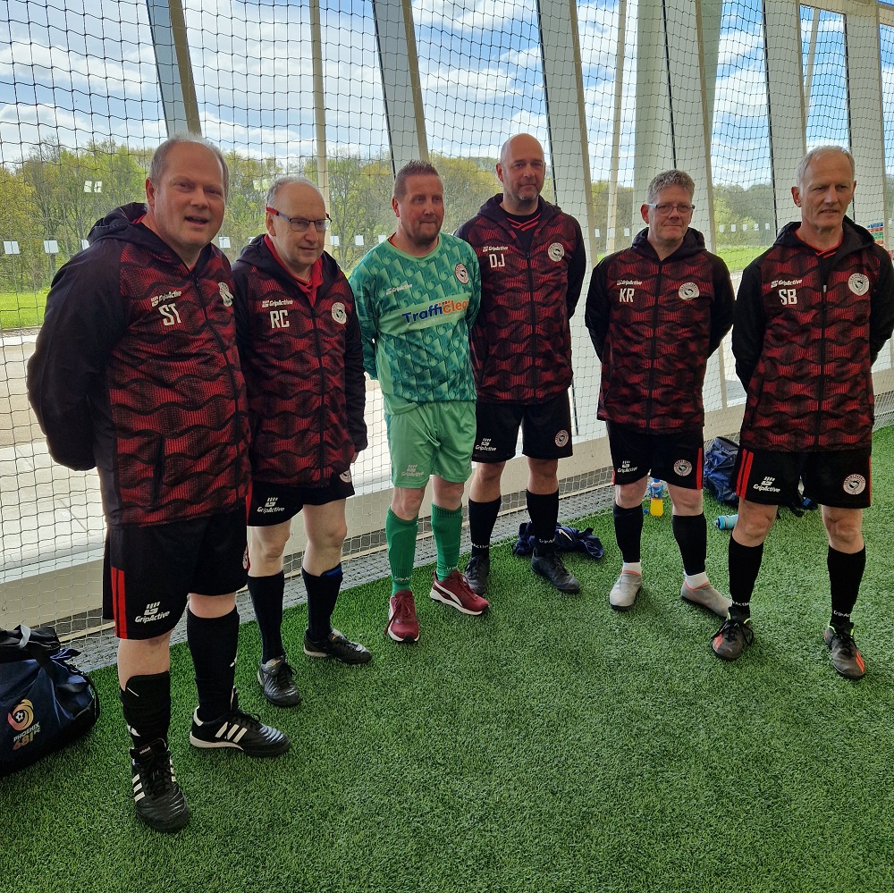 A walking football team posing together indoors beside a net, with six players in matching black and red patterned kits, except for one in a green goalkeepers jersey. They're standing on artificial grass with a hint of natural light streaming in, suggesting an indoors sports facility. 
