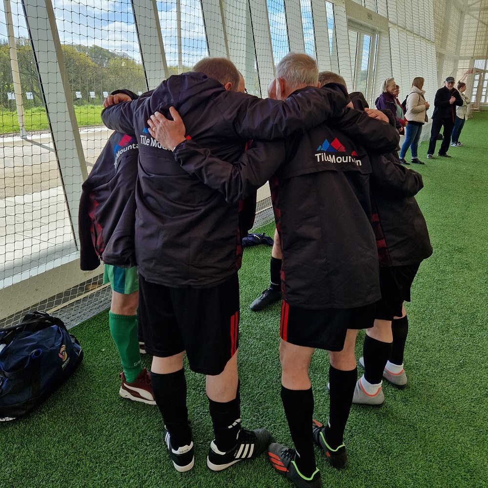 Members of a walking football team in a huddle, viewed from behind, showcasing their black jackets with red accents and Tile Mountain logo on the back. The team is standing on a green artificial turf field insides a brightly lit sports facility. 