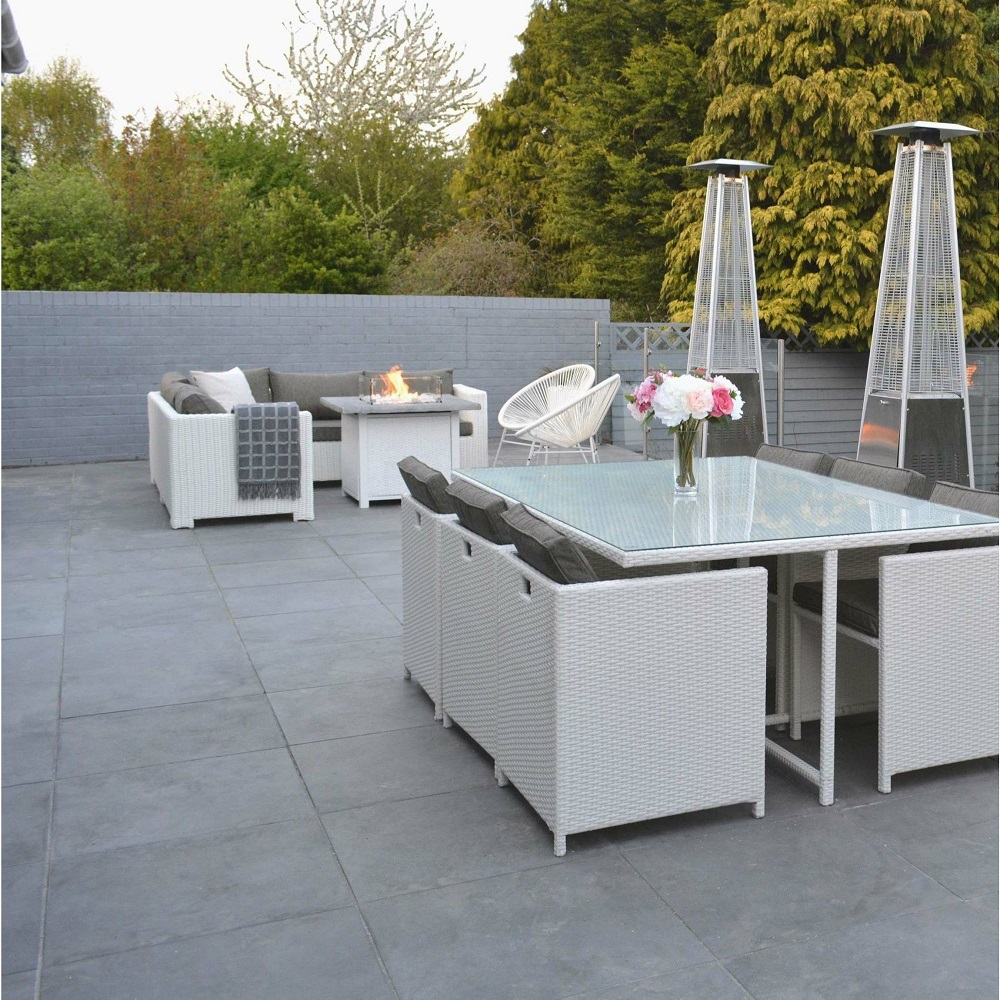 A modern outdoor dining space featuring dark grey square slab tiles that create a sleek and stylish foundation. The are is furnishes with white wicker dining chairs and a matching table with a glass top, accented by soft grey cushions. A cosy lounge area with a sofa and fire pit table adds warmth to the space, flanked by elegant glass tube patio heaters. Surrounded by a brick walls and greenery, this patio merges comfort with contemporary design, perfect for entertaining or relaxing evenings. 
