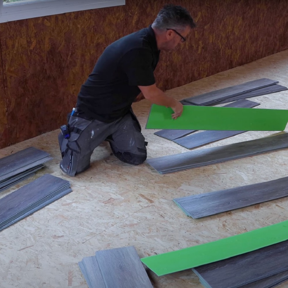 Man kneeling on a wooden subfloor while arranging planks of varying grey tones for flooring installation, ensuring even distribution of colour.