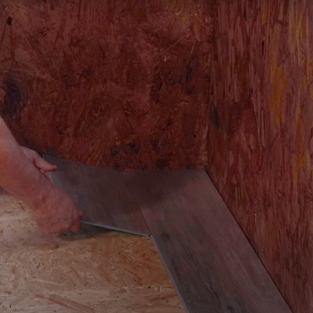 The image shows a close up of hands carefully placing a laminate flooring plank at the start of the second row against the wall, ensuring proper alignment and fit for the continuation of the floor installation. 