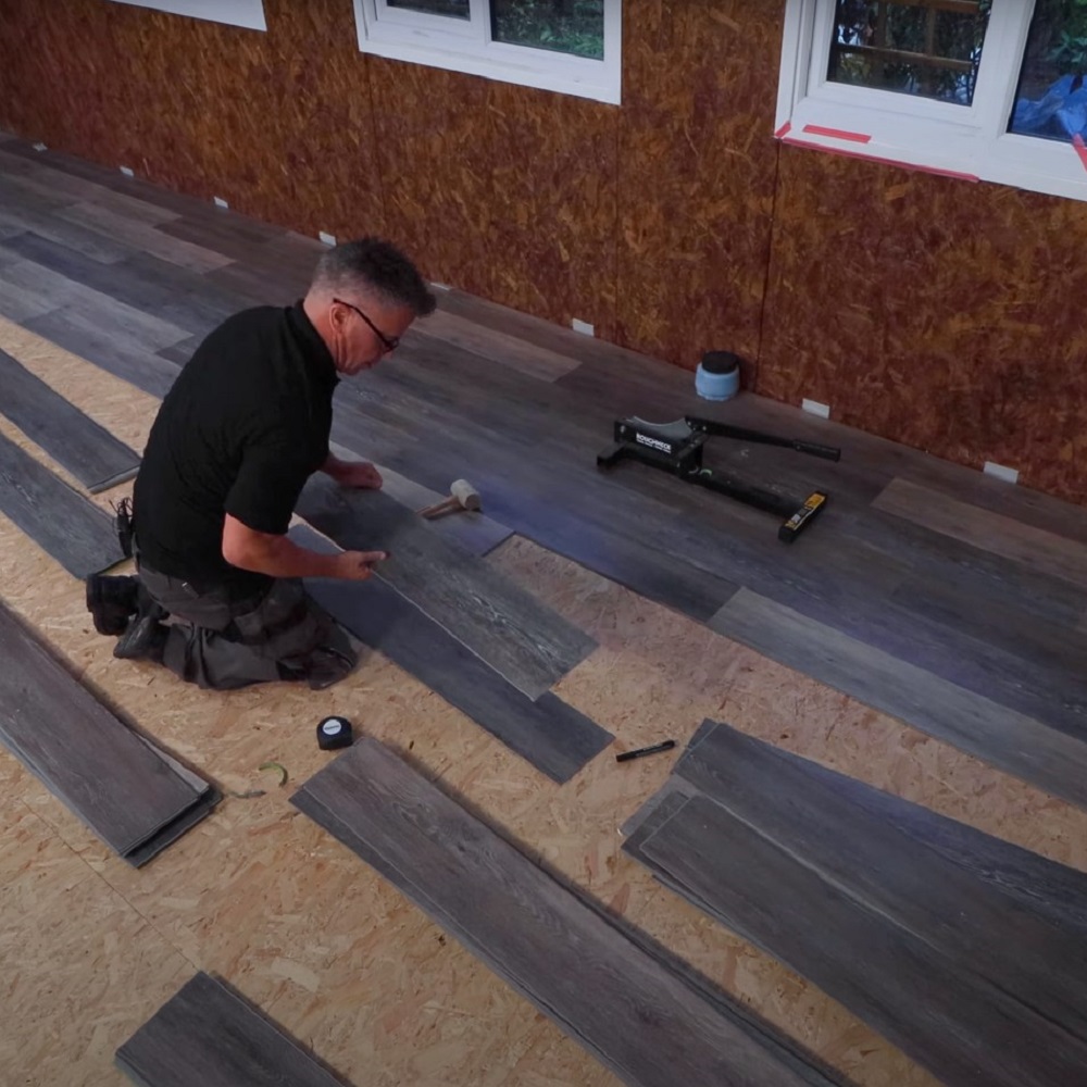 A man kneeling on a subfloor surrounded by various shaded grey LVT planks mid-installation. They have a rubber mallet to snuggly fit the planks together. 