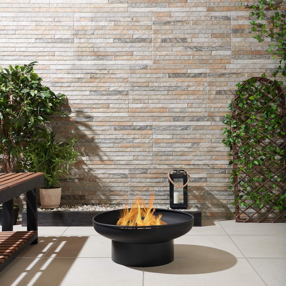 A cosy outdoor corner featuring a natural split face wall tile in neutral tones, complemented by a modern black fire pit in the centre. The area is accented with greenery, including potted plants and ivy climbing a trellis, creating a serene space for relaxation and entertainment. The floor tiles are a light sandy colour which beautifully contrasts the textured wall and the sleek fire feature. 