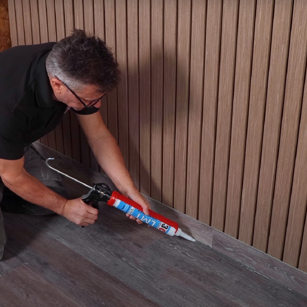 In the image, a man is shown applying silicone sealant along the edge where an LVT floor meets the vertical wall panelling, using a caulk gun for a precise finish as part of the final steps in floor installation. 