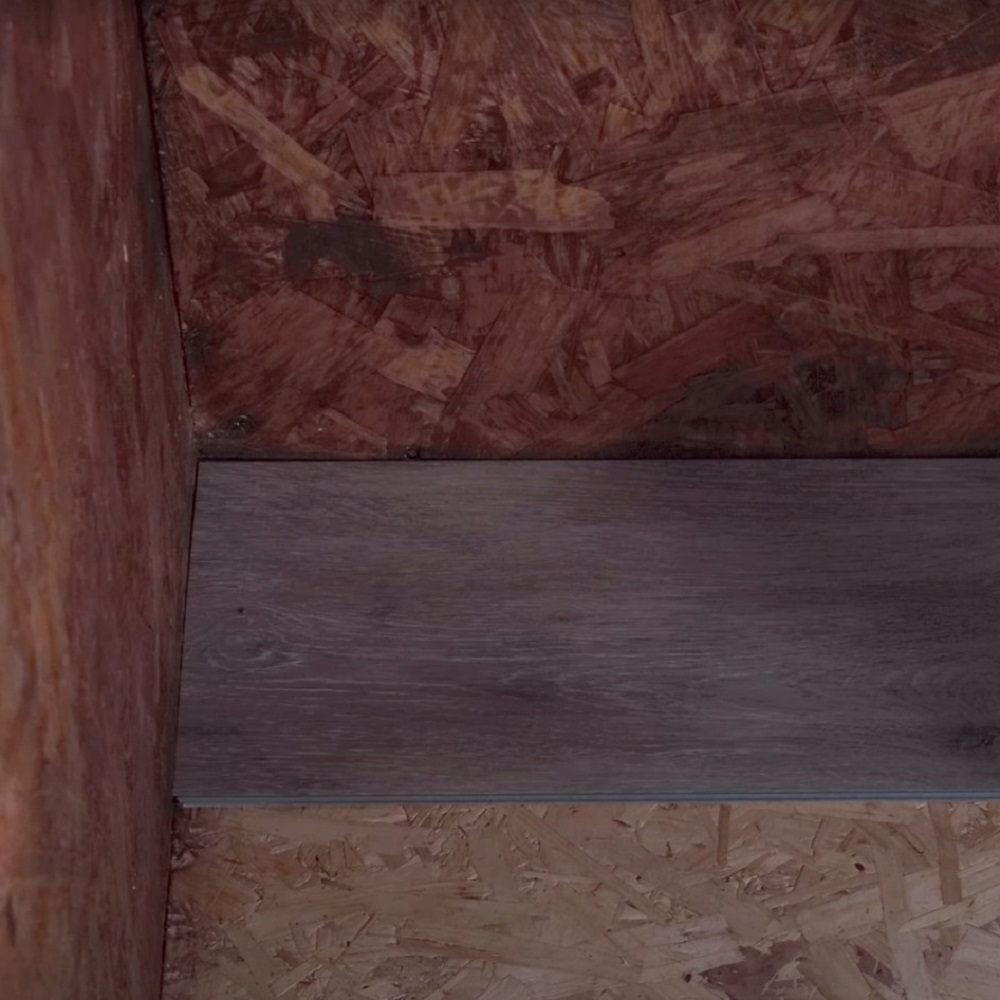 The image shows a close up view of a corner of a room where the first plank of grey LVT flooring has been laid down on a wooden chipboard subfloor, marking the beginning of a flooring installation. 