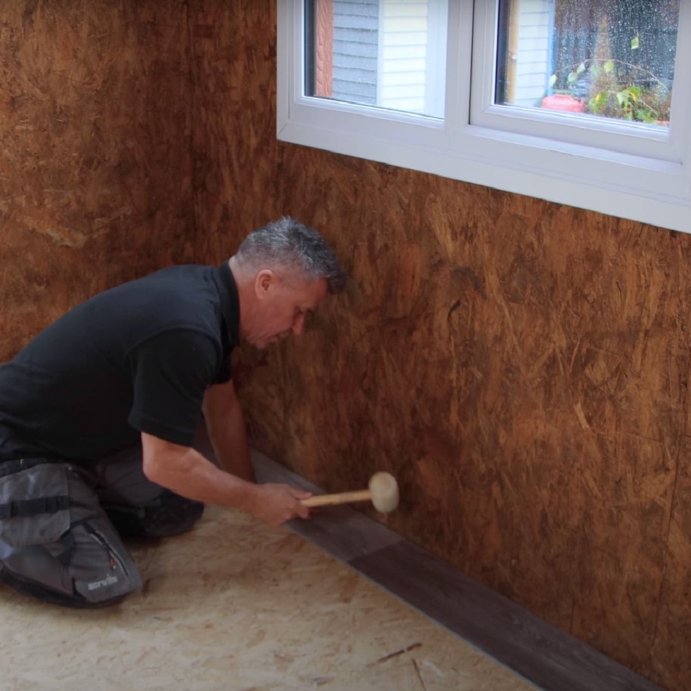 Man kneeling on the floor, gently tapping a dark LVT floor plank into place with a rubber mallet near a windowed wall with a wood finish. 