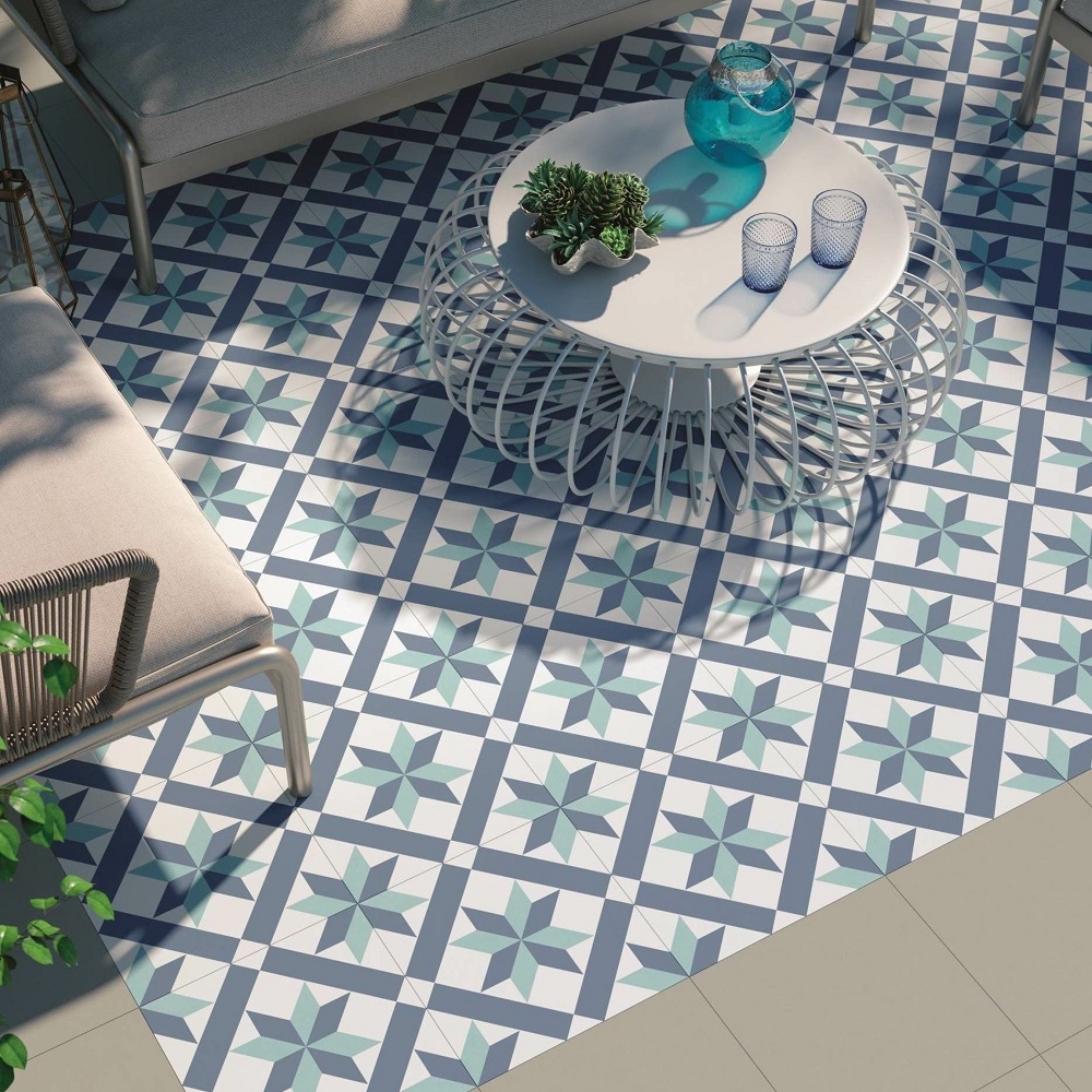 A vibrant outdoor setting with eye-catching patterned porcelain tiles in shades of blue and white, arranged in a starburst design. The scene features a chic round metal wire table atop the tiles, adorned with a small potted plant and decorative items, including a glass vase and blue drinking glasses, Part of a grey outdoor sofa and lush plants are visible in the periphery, all bathed in natural sunlight, evoking a fresh and inviting atmosphere perfect for a relaxing outdoor retreat. 