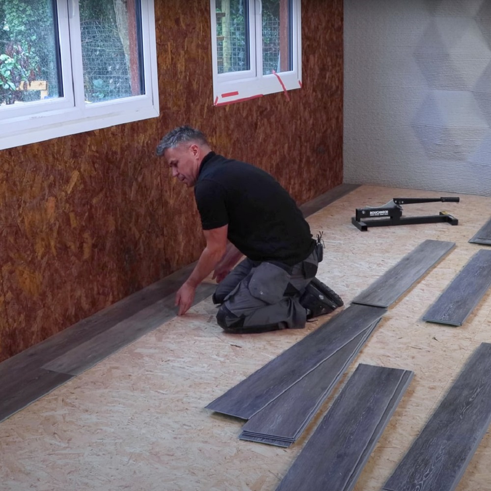 A man kneeling on a wooden subfloor, actively laying grey LVT planks in the second row of a room that features a windowed wall. Flooring tools visible in the background, indicating an ongoing installation process. 