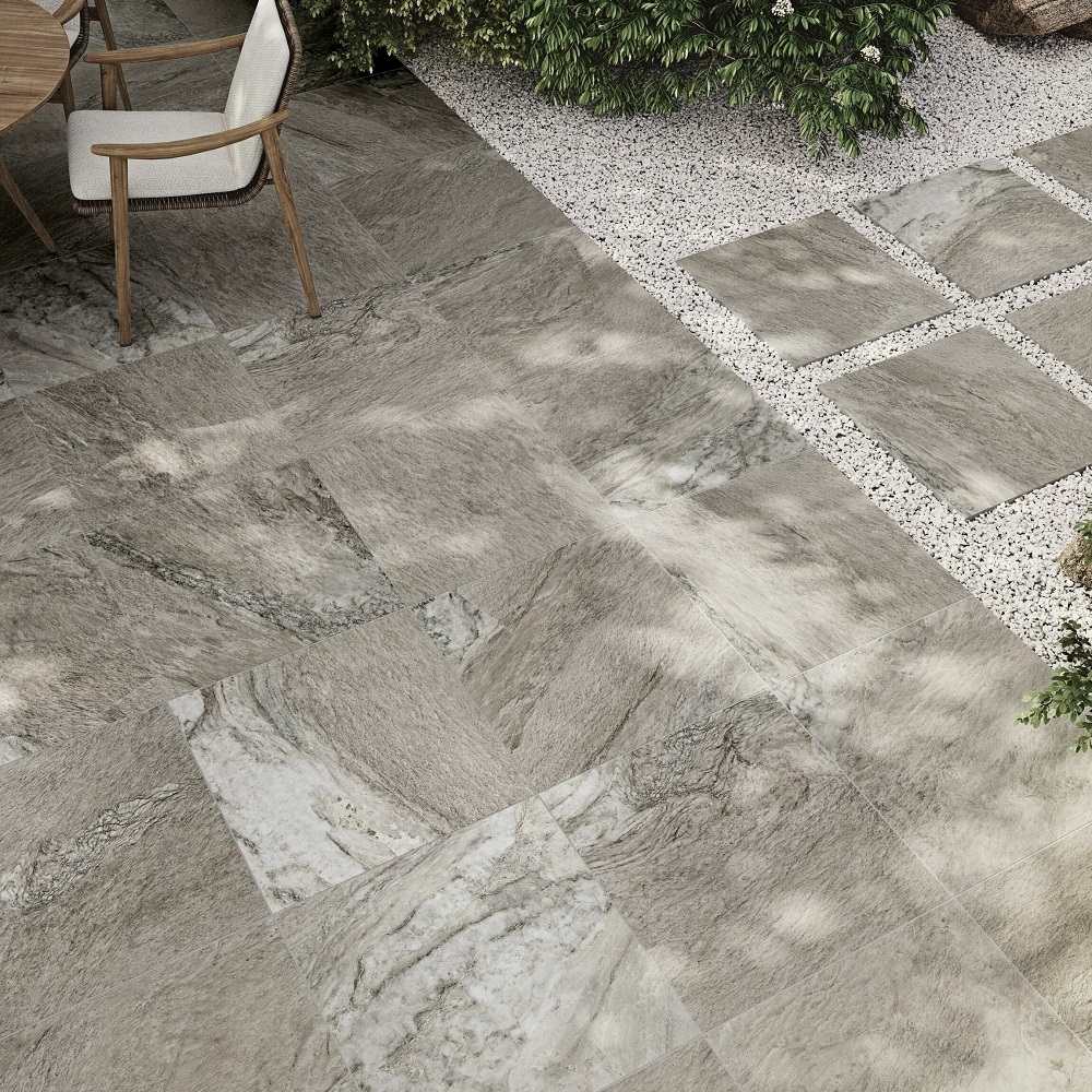 Outdoor patio featuring grey stone effect matt porcelain tiles in a varied pattern, bordered by small white pebbles and lush green outdoor plants. 