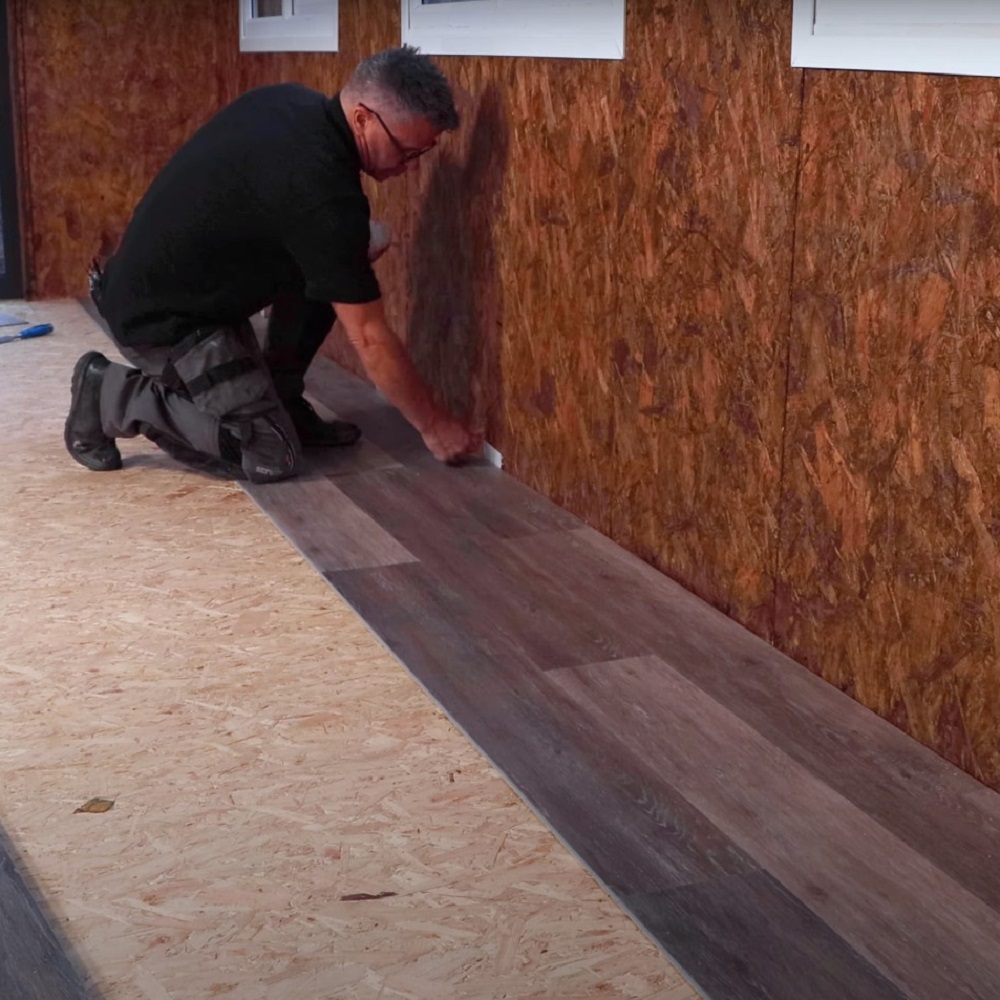 A man kneeling and placing spacers along the wall to maintain a gap while installing a row of dark LVT flooring, ensuring the planks are correctly aligned against a wall with windows. 