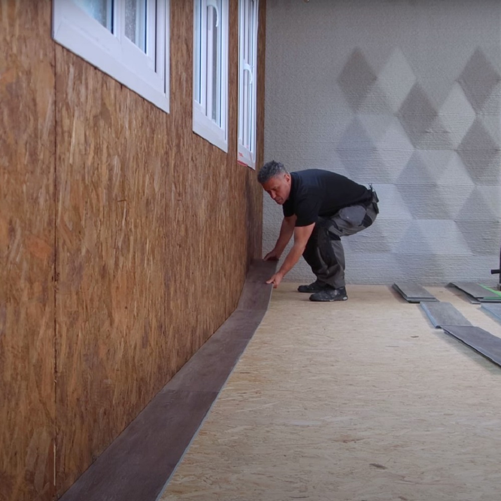A man crouched over, carefully placing a long, dark plank of LVT flooring along the first row adjacent to a wall, under a set of windows. The room is partially completed with insulation visible on the opposing wall. 