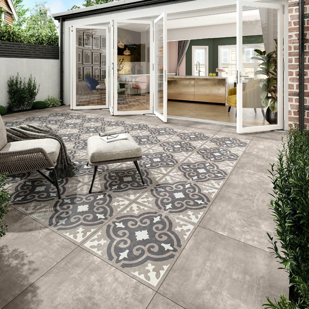 An elegant outdoor patio featuring grey concrete effect matt porcelain slab tiles with a central decorative pattern in shades of grey, blue and white. Wicker lounge chairs with soft grey cushions and a matching ottoman invite relaxation, complementing the open bi-fold doors that lead to a stylish interior. Lush green shrubbery borders the space, enhancing the indoor outdoor flow and creating a serene environment. 