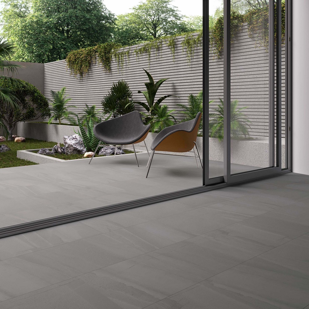 A contemporary outdoor area with dark grey porcelain floor tiles leading to a tranquil garden corner, adorned with sleek modern chairs, lush tropical plants, and a sculptural wall feature, all visible through floor to ceiling glass doors. 