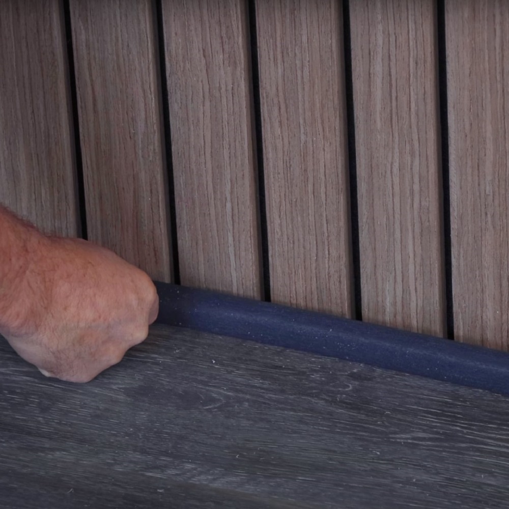 A close-up of a hand pressing a dark scotia beading into place along the junction where the grey laminate flooring meets the vertical wood panelling, covering the expansion gap as part of the flooring installation process. 