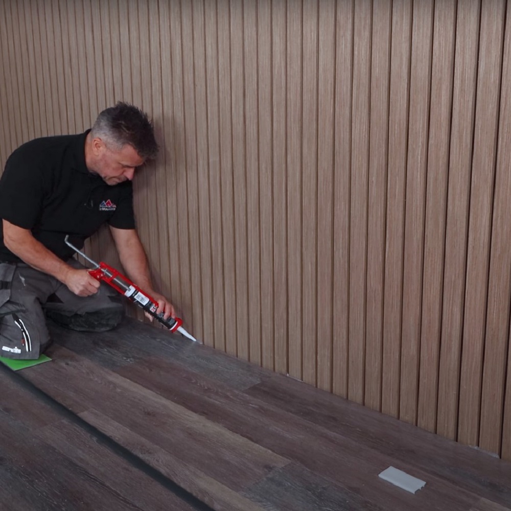 The image shows a person kneeling and applying sealant to the edge of LVT floor plank. They are focused on the task, with a caulk gun in hand against a backdrop of vertical wood panelling. 