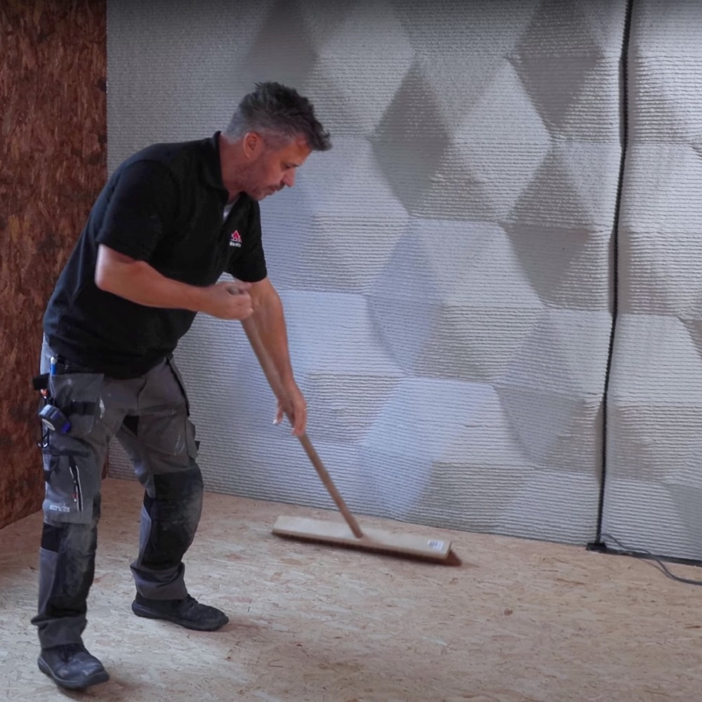 A man sweeping a wooden subfloor with a wide brush, preparing the surface for flooring. Behind them a textured wall insulation creates geometric patterned backdrop. 