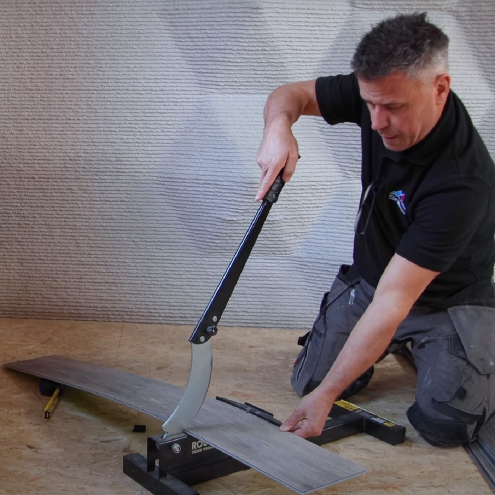 A man is shown using a laminate flooring cutter to trim a grey plank to size. They are focused on the task, pressing down on the cutters handle to make a precise cut, with the backdrop of an insulated wall behind them. 